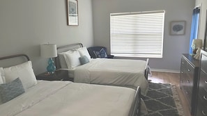A bedroom with two beds, a nightstand with a lamp, a dresser, and a window with blinds.