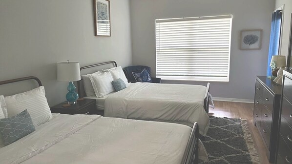 Quarto com duas camas, mesa de cabeceira com abajur, cômoda e janela com persianas.