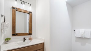 A bathroom with a wooden vanity, a white sink, a mirror with lights, a towel rack, and a small plant. - Cozy Cabin with Private Hot Tub, Fireplace & Community Amenities Near Thunder Ridge & Table Rock (Hollister)