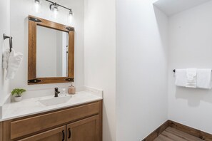 Un baño con tocador de madera, un lavabo blanco, un espejo con luces, un perchero para toallas y una planta pequeña.