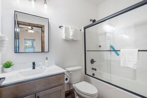 Un baño moderno con cabina de ducha de vidrio, inodoro blanco, lavamanos con espejo y una pequeña planta en maceta sobre la encimera.