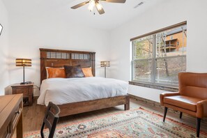 Un dormitorio con una cama de madera, un ventanal grande, un ventilador de techo y un sillón de cuero.