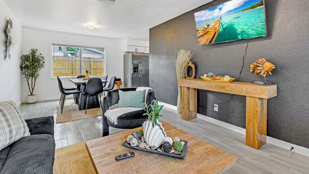 A modern living room with a TV mounted on the wall, a dining area with a table and chairs, and a kitchen in the background.