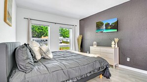 A bedroom with a bed, a dresser, a painting, and a view of greenery outside.