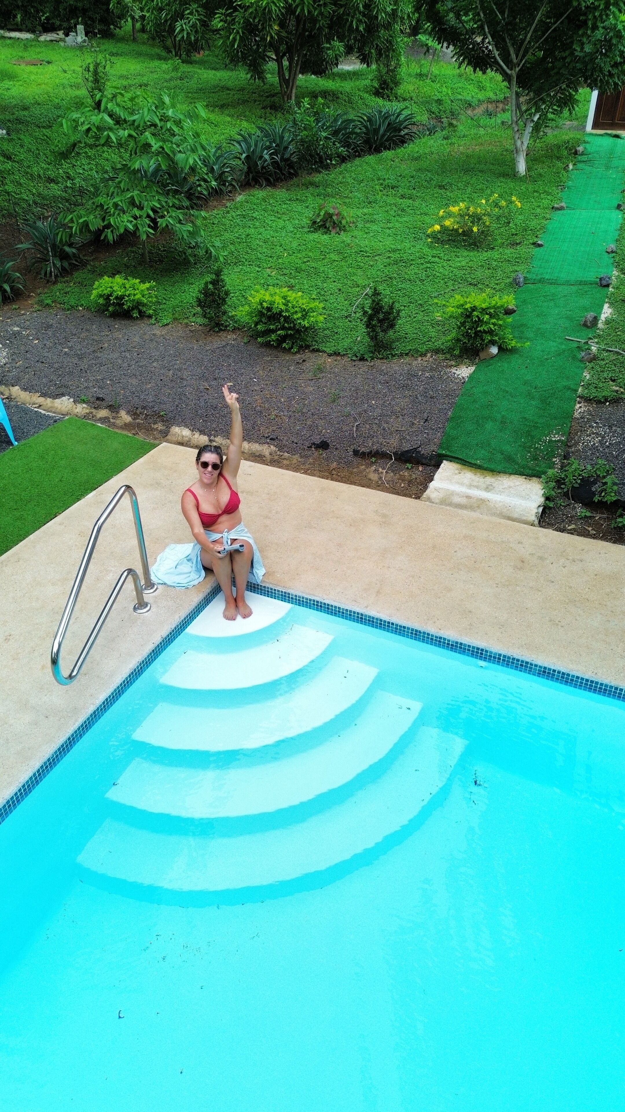 A person sitting by a swimming pool with a green lawn and trees in the background.
