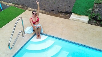 A person sitting by a swimming pool with a green lawn and trees in the background.