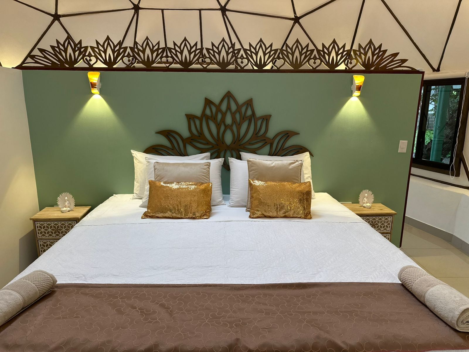 A neatly made bed with white linens and two gold pillows, a decorative wall headboard, and bedside tables with small figurines.