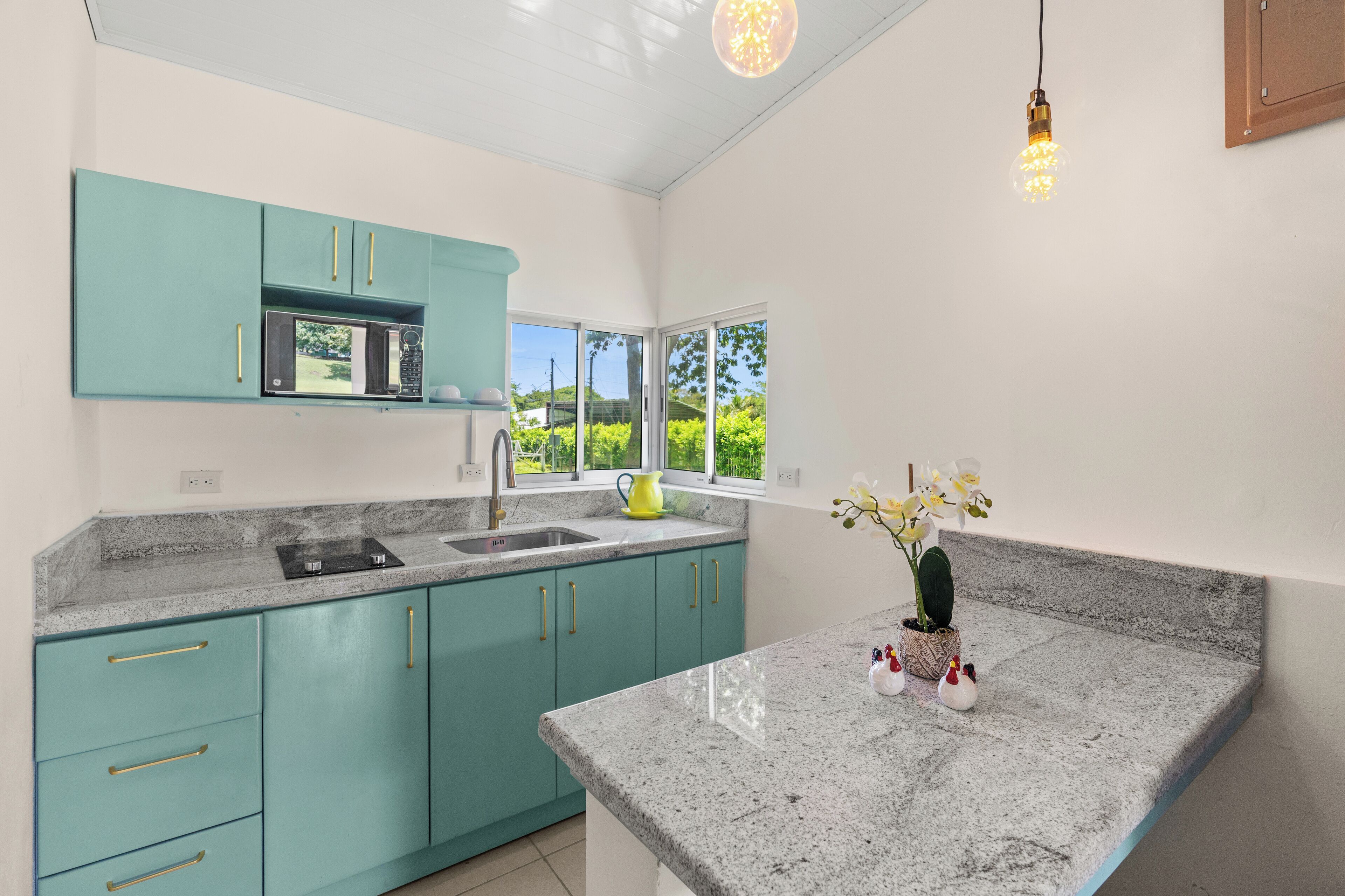 A kitchen with teal cabinets, a granite countertop, a microwave, and a window with a view of greenery.