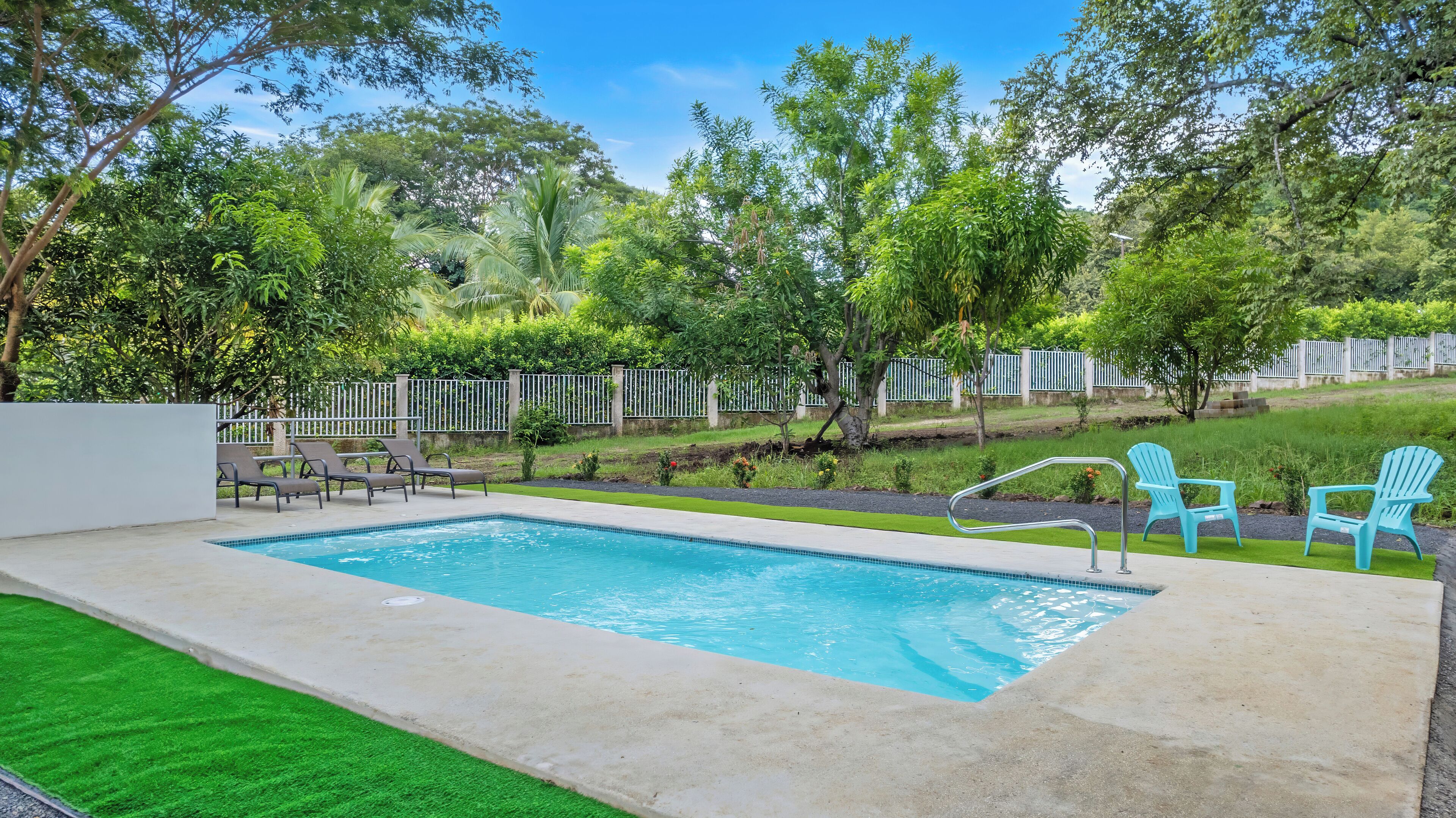A rectangular swimming pool with a tiled edge, surrounded by a concrete deck. There are four lounge chairs and two blue Adirondack chairs.