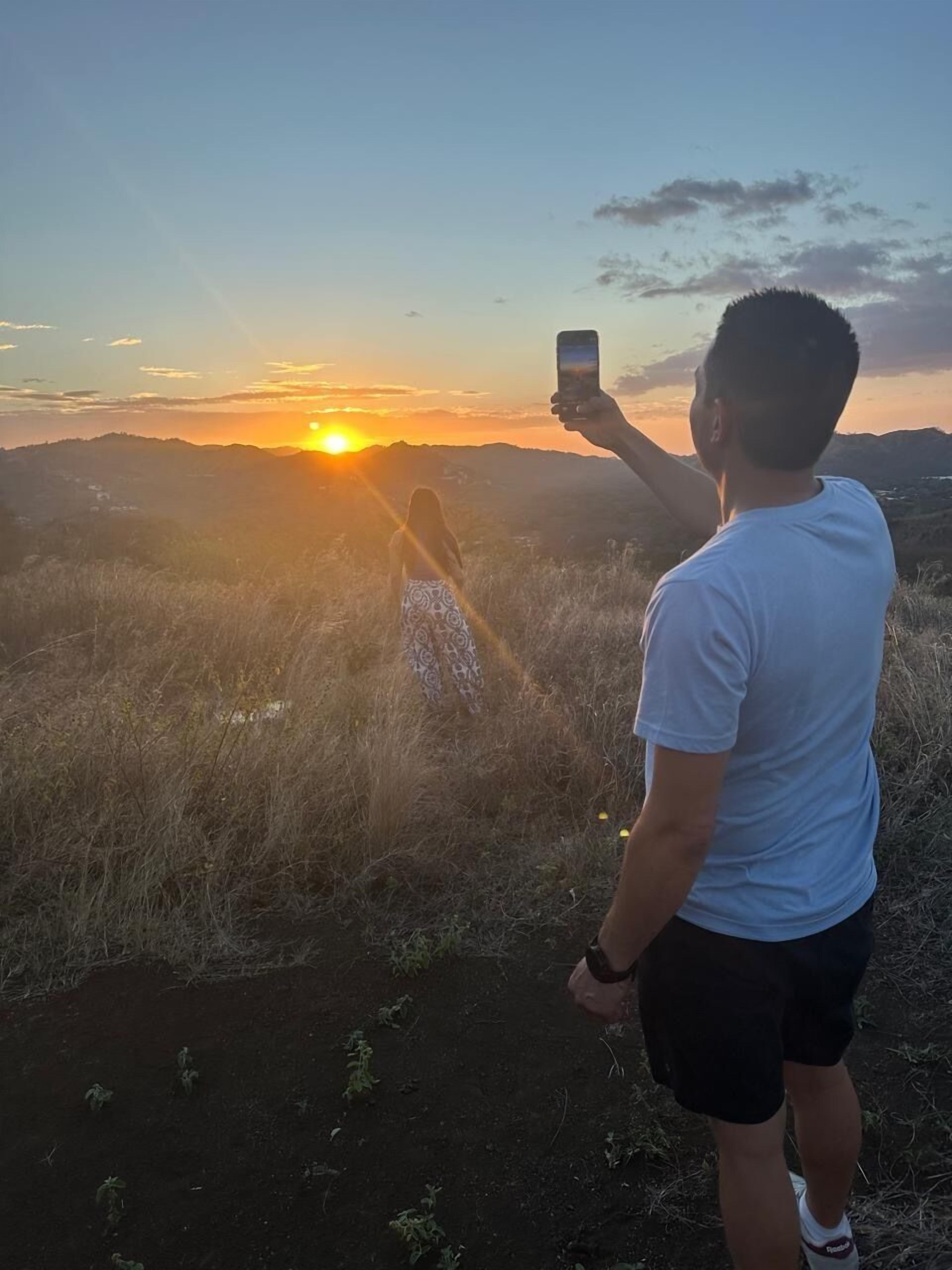 Une personne prend une photo d’un coucher de soleil, avec un paysage en arrière-plan.
