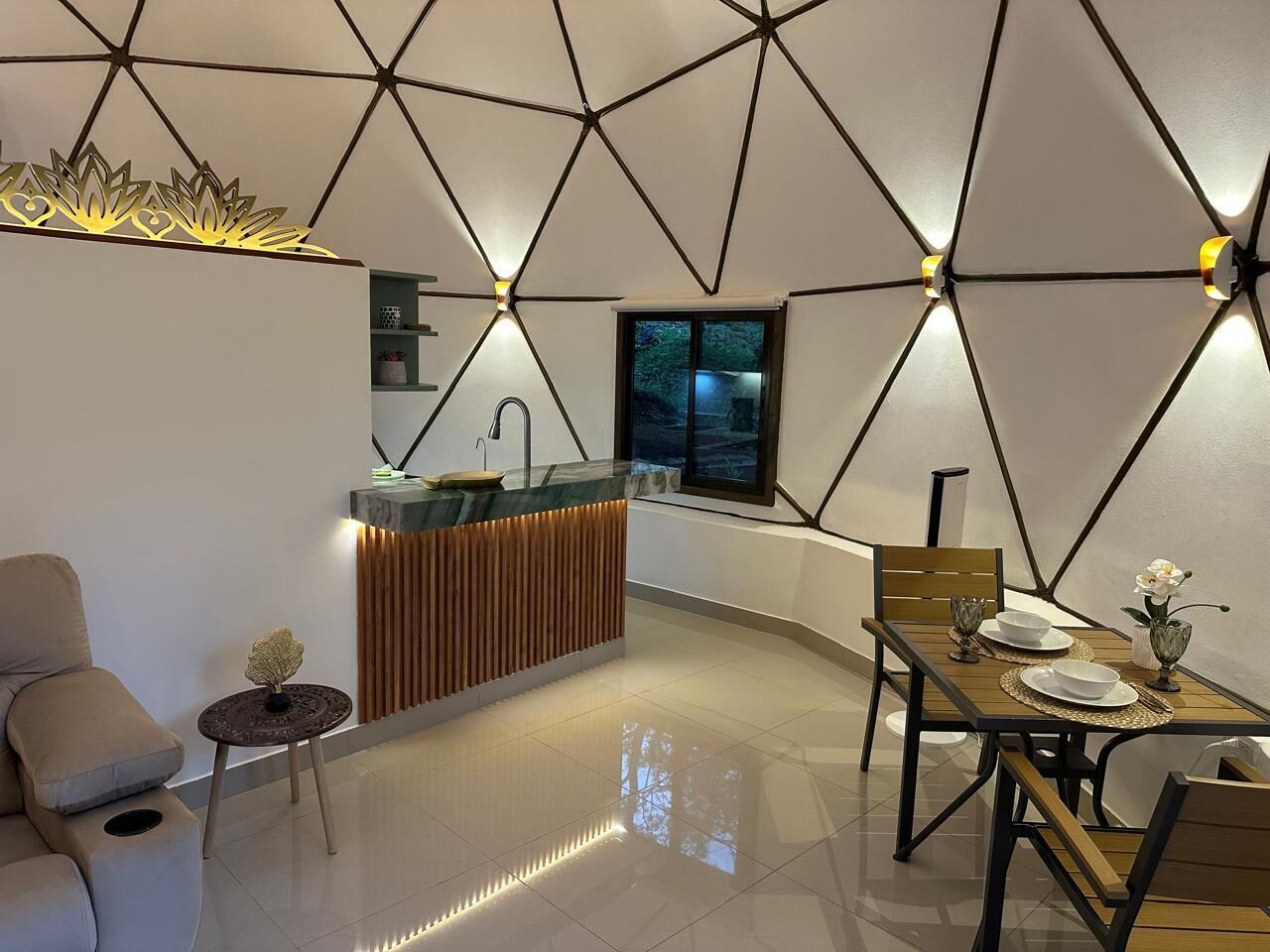 Een modern interieur met een geometrisch plafondontwerp, een eethoek met een houten tafel en stoelen, een keuken met een inbouw kookeiland en een zithoek met een bank en een bijzettafeltje.