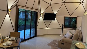 Un intérieur moderne avec un plafond au design géométrique, un téléviseur à écran plat fixé au mur, un coin repas avec une table et des chaises, et un canapé offrant une vue sur l’extérieur.