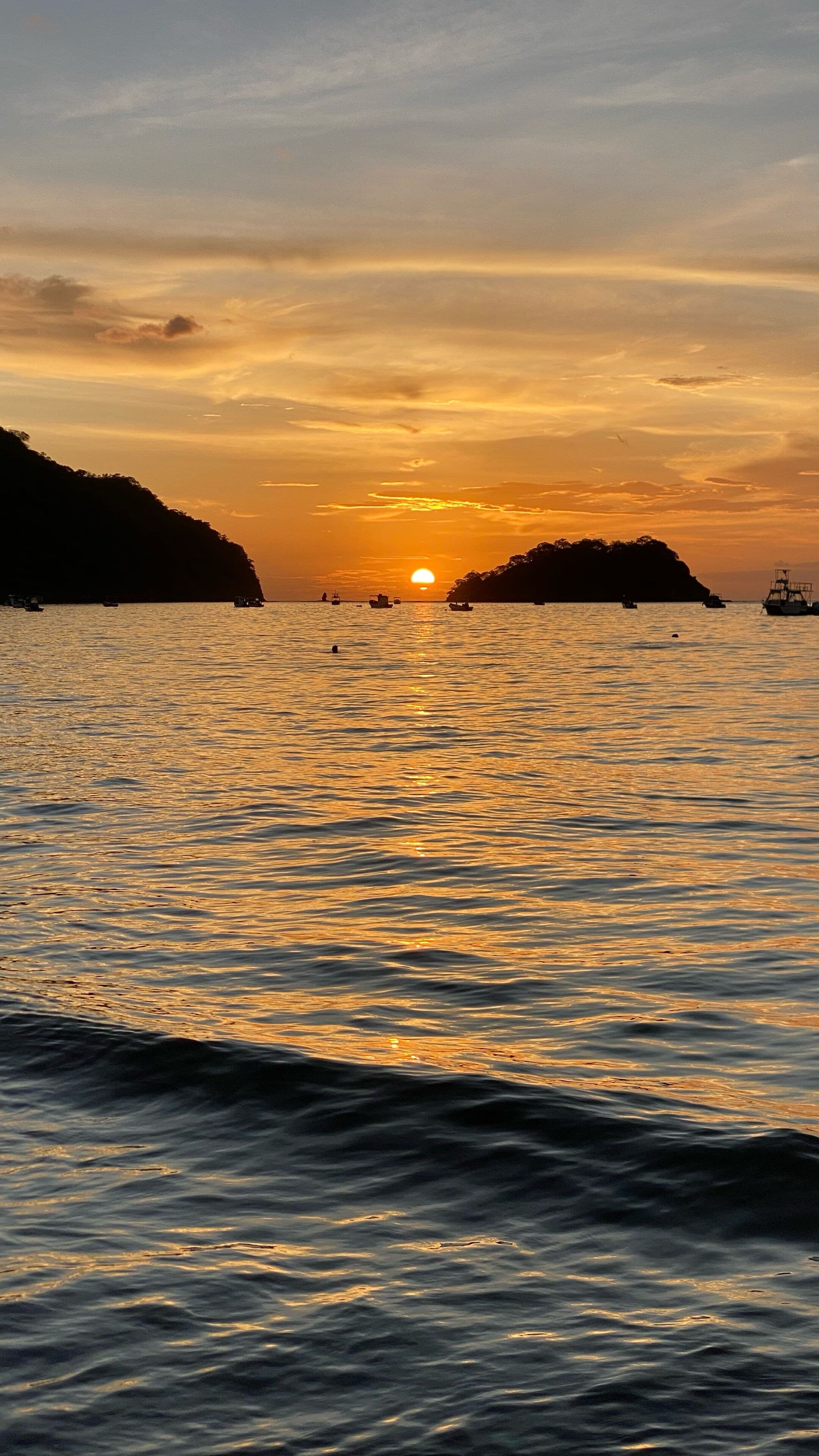A sunset over a calm sea with boats and distant landforms.
