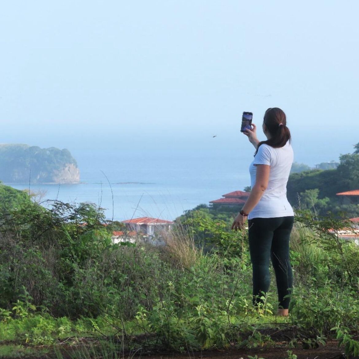 A person taking a photo with a smartphone, overlooking a coastal area with vegetation and buildings in the background.