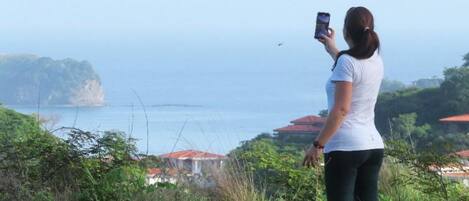 A person taking a photo with a smartphone, overlooking a coastal area with vegetation and buildings in the background.