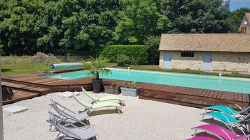 Una piscina con cubierta de madera y sillas de descanso, rodeada de follaje y un edificio de piedra.
