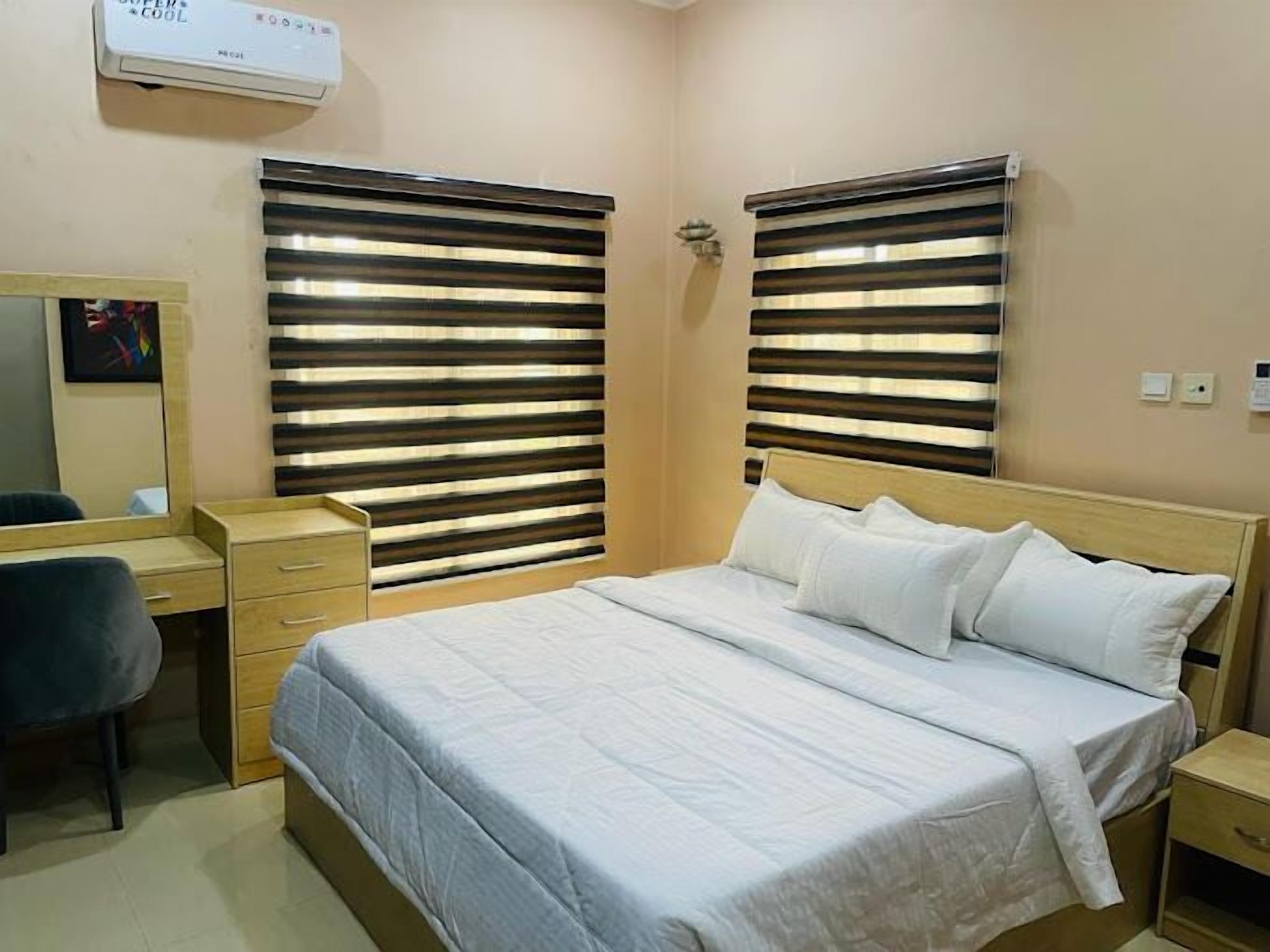 A bedroom with a bed, bedside tables, a desk, and a chair. There are two windows with horizontal blinds.