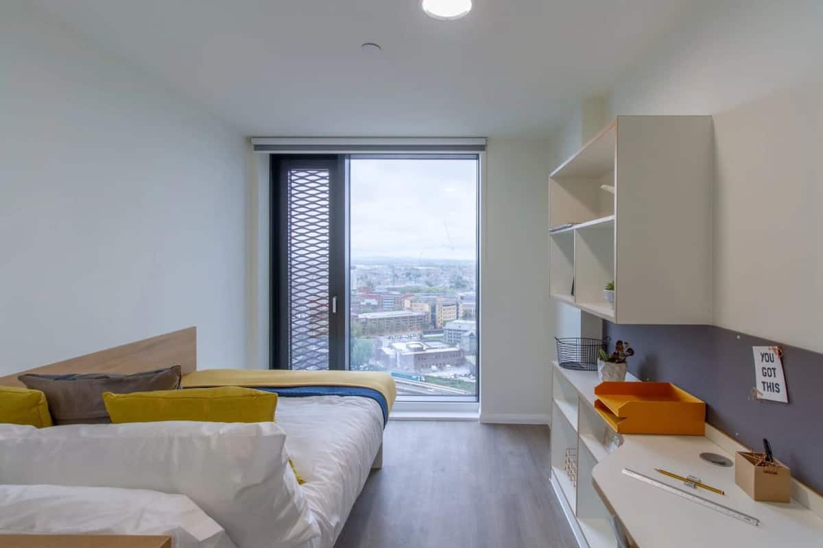 A modern bedroom with a bed, a desk, and a view of the city through a large window.