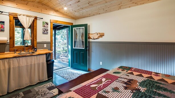 Quarto com teto de madeira, cama com colcha, janela com cortinas e porta que dá para o exterior.