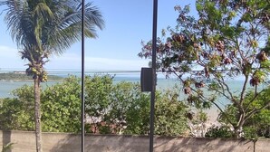 Una habitación con un amplio ventanal que da a una playa, palmeras y un coche estacionado afuera.