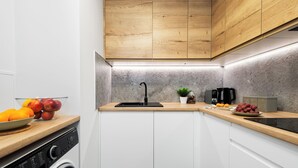 Une cuisine compacte équipée d’un lave-linge, de meubles en bois, d’un évier et d’un plan de travail sur lequel on trouve des fruits et une machine à café.