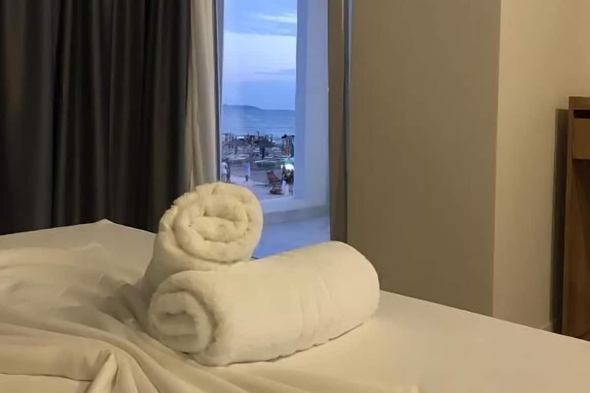 A hotel room with a bed, a window with a view of a beach and people, and a towel on the bed.