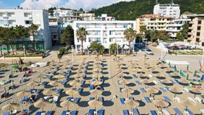 A beach with numerous sun loungers and umbrellas, surrounded by buildings and palm trees. - Hotel Besani (Durrës)