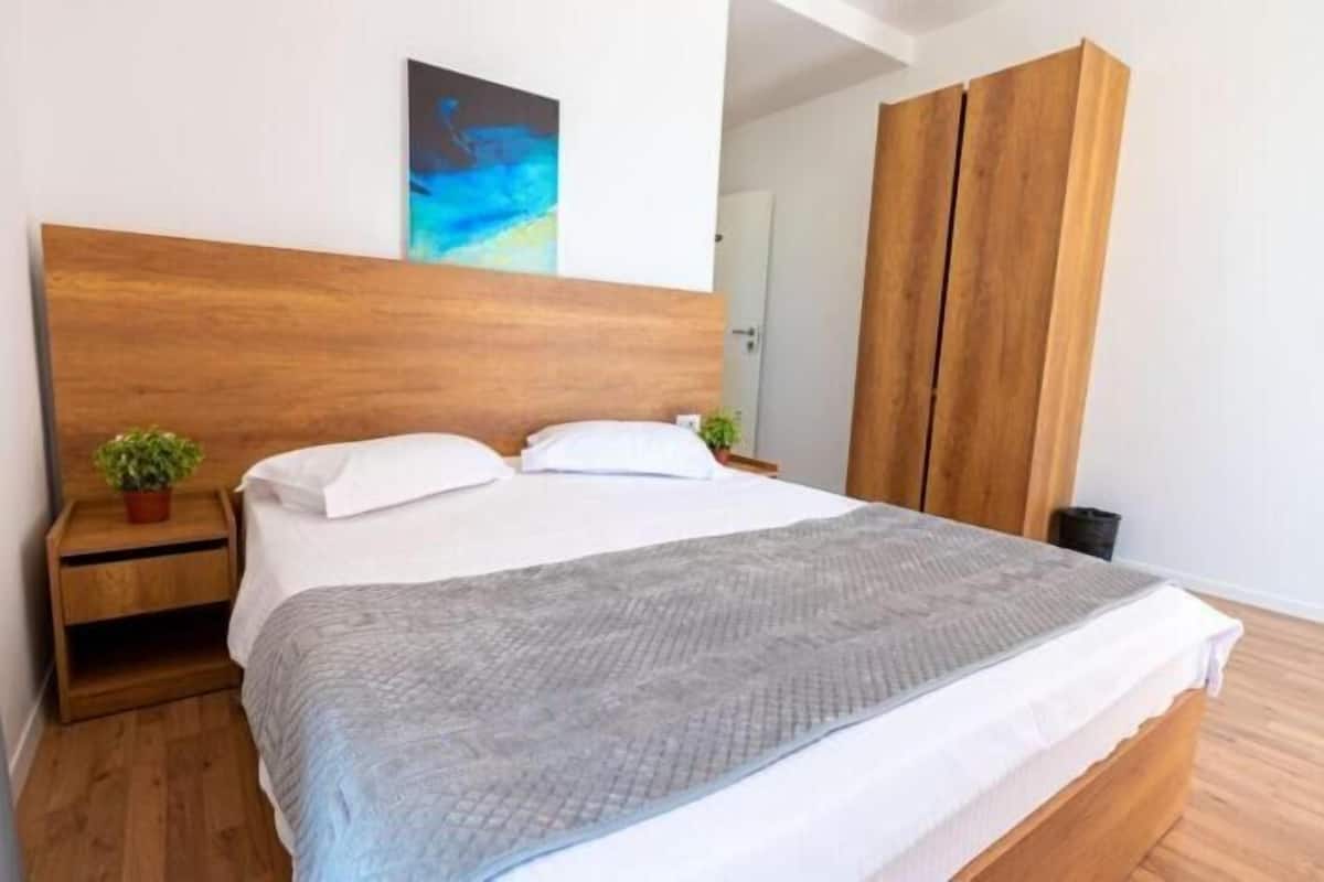 A modern bedroom with a wooden headboard, a bed with white linens, a grey throw blanket, a small bedside table with a plant, and a wardrobe.