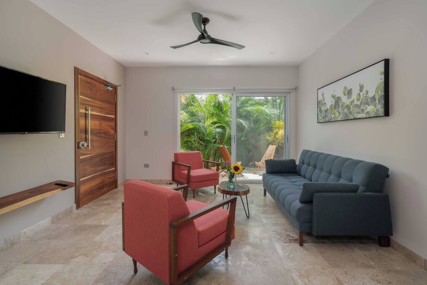 A modern living room with a flat-screen TV, a ceiling fan, a large window with a view of greenery, a sofa, and two armchairs.