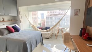 A modern bedroom with a bed, a hammock, a small table, and a view of the city. - Condomínio ver de Panorama (Sao Paulo)