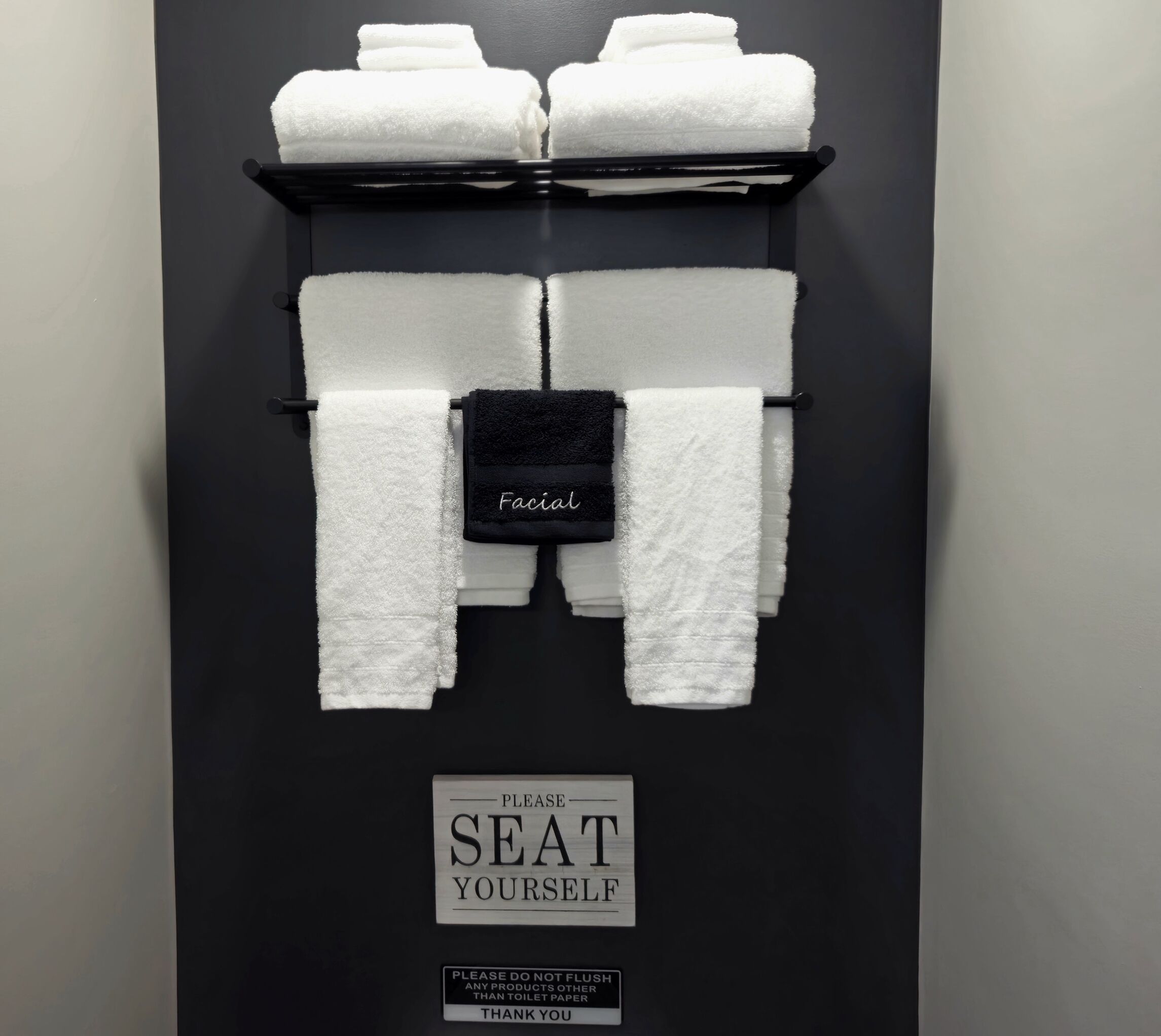 A bathroom with a towel rack holding multiple white towels, a black towel with "Facial" written on it, and signs instructing to "Please Seat Yourself" and "Please do not flush any products other than toilet paper."