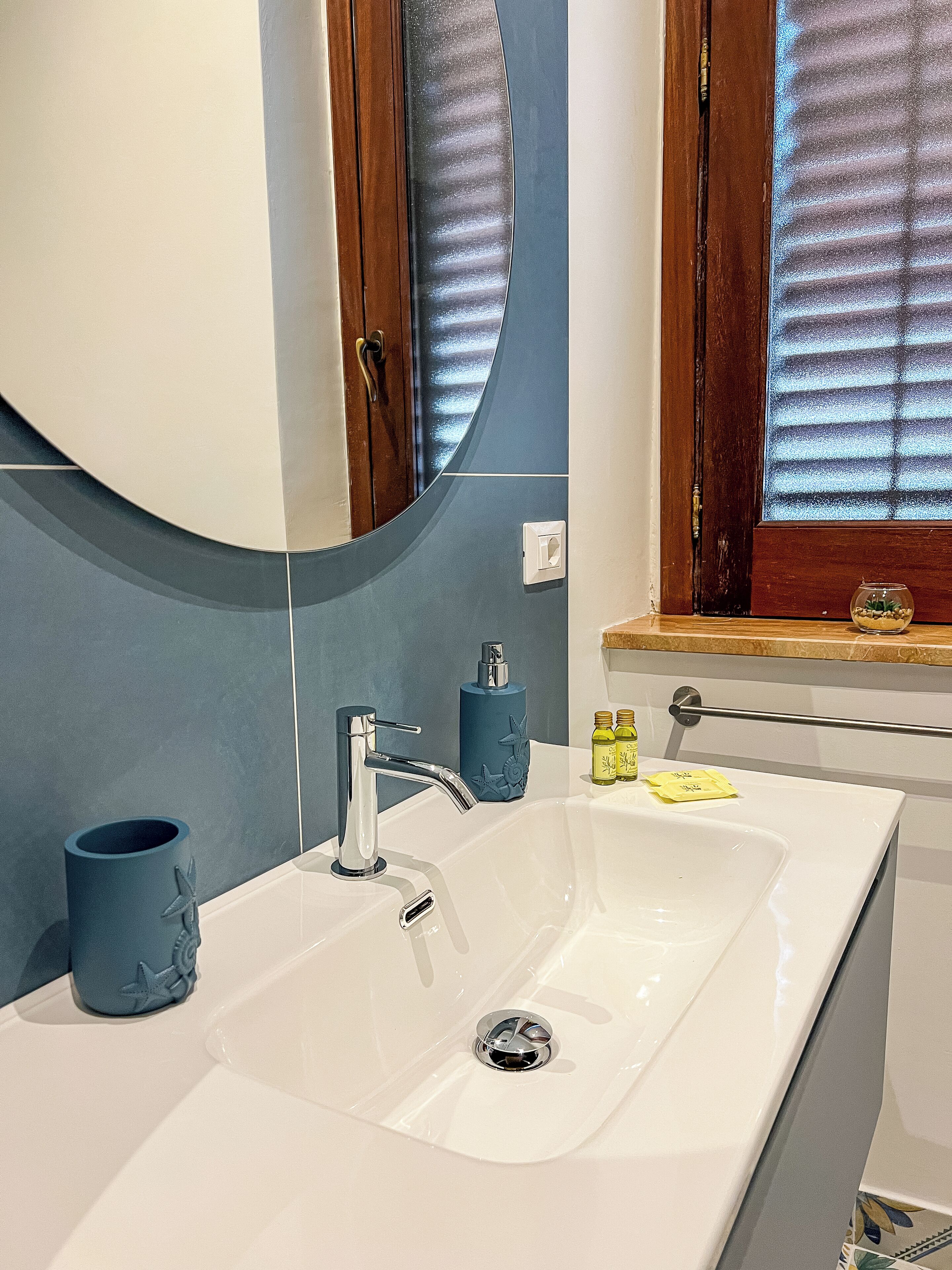 Un bagno con un lavandino bianco, uno specchio, una finestra con persiane e una porta in legno.