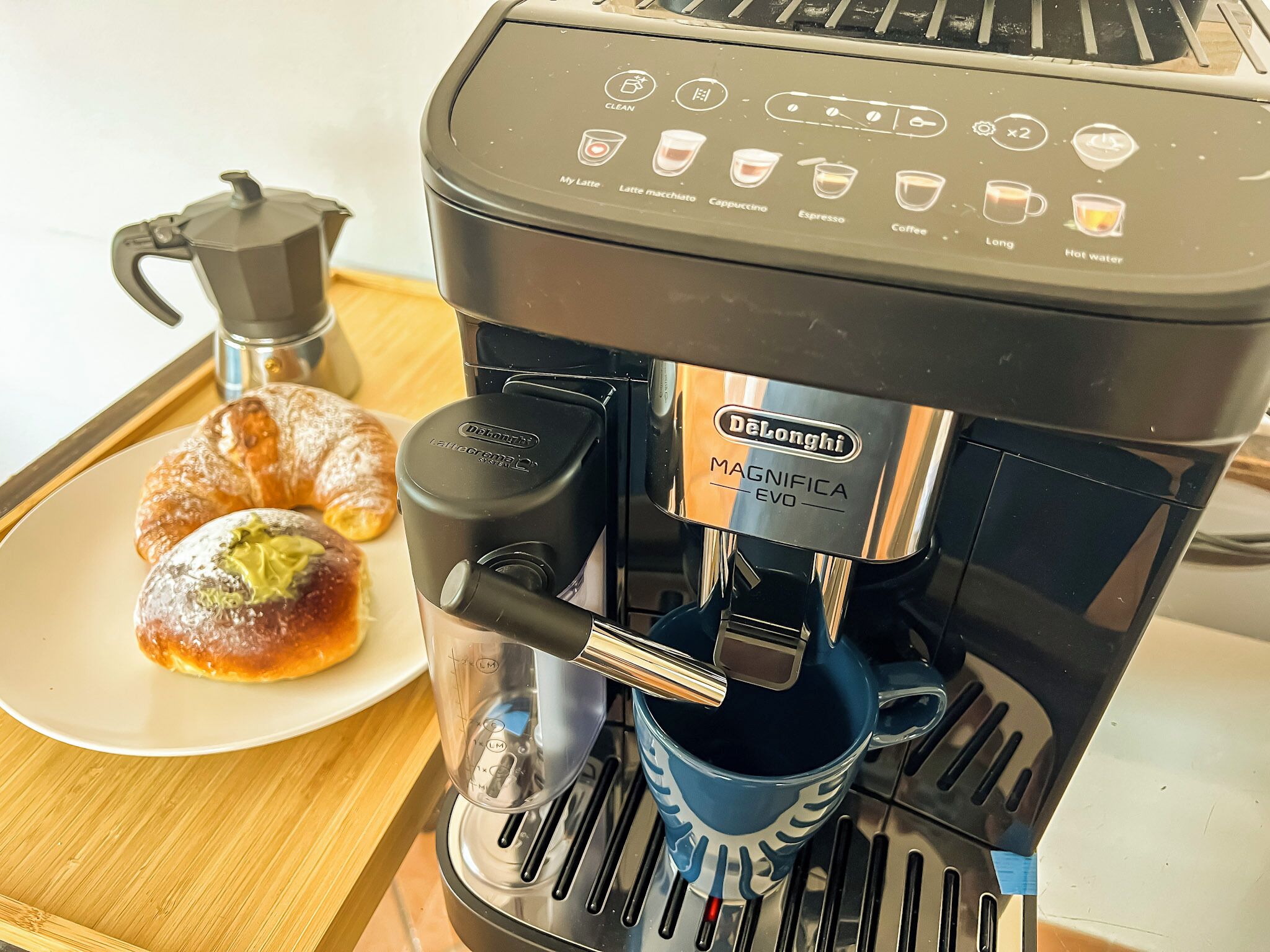 A DeLonghi Magnifica EVO coffee machine with a coffee pot and a plate of pastries on a tray.