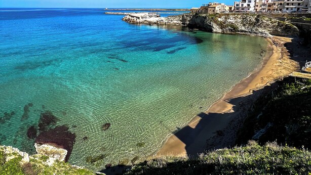 Blick auf die Küste mit klarem türkisfarbenem Wasser, einem Sandstrand und Gebäuden entlang der Uferlinie.