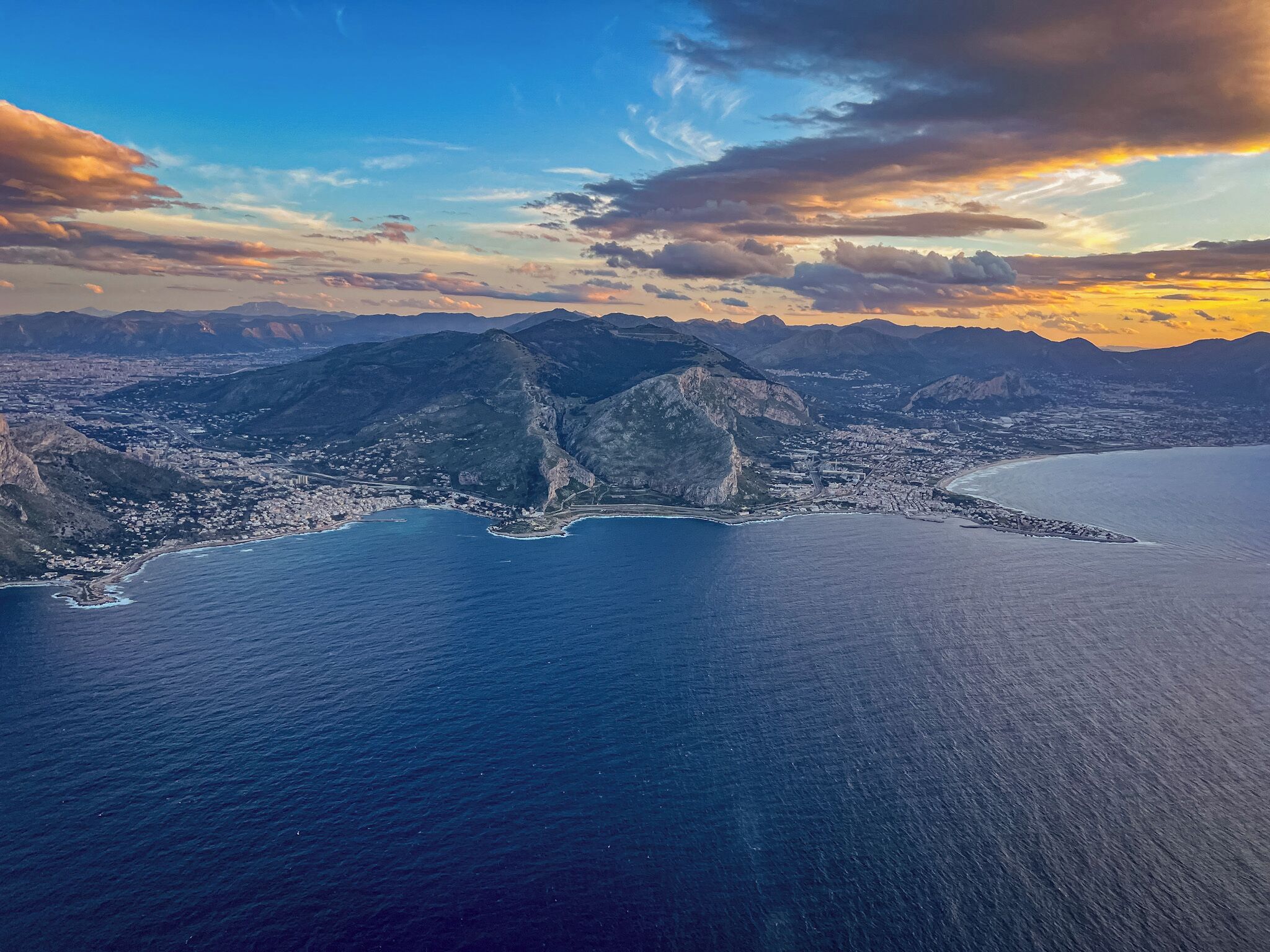 Une vue aérienne d’une ville côtière, avec une chaîne de montagnes imposante, une baie et un ciel dégagé.