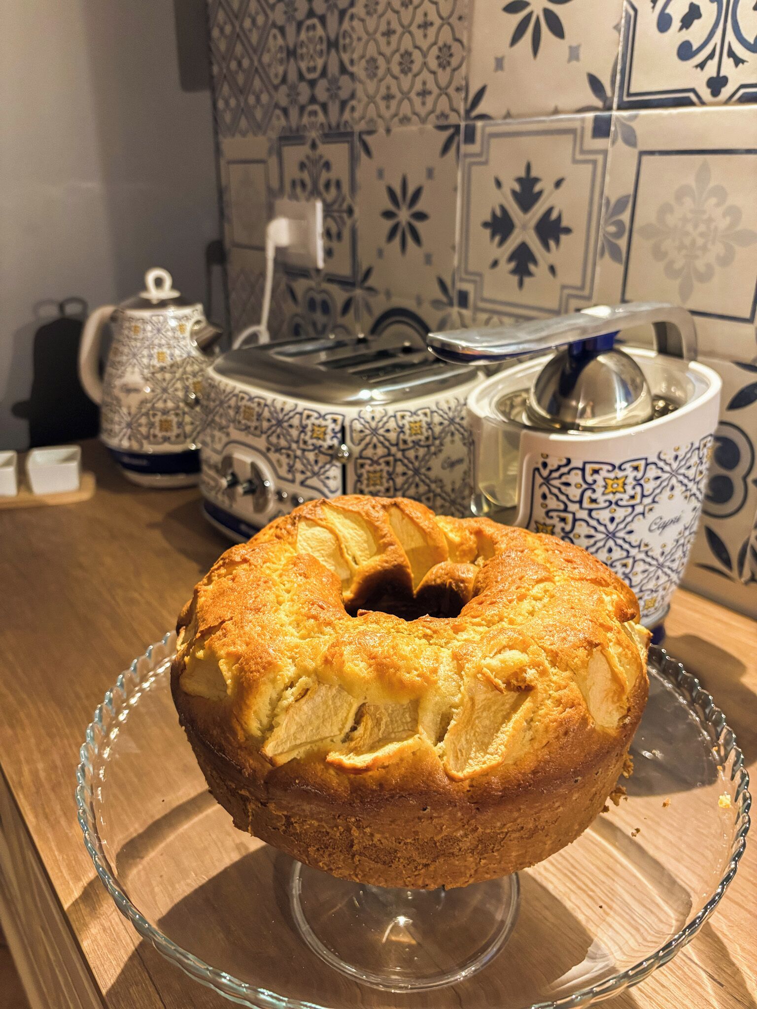 A cake on a glass plate with a patterned ceramic mug and a teapot in the background.