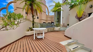 A modern outdoor patio with a round table and benches, surrounded by tropical plants and a stone wall. - Cobertura Triplex no Guarujá SP 1 (Guaruja)