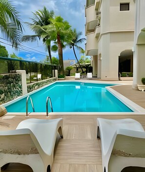 Une piscine entourée de chaises longues, de palmiers et d’un bâtiment doté de balcons.