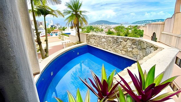 A swimming pool with a stone wall and tropical plants, overlooking a coastal town with buildings and mountains in the distance. - Cobertura Triplex no Guarujá SP 1 (Guaruja)