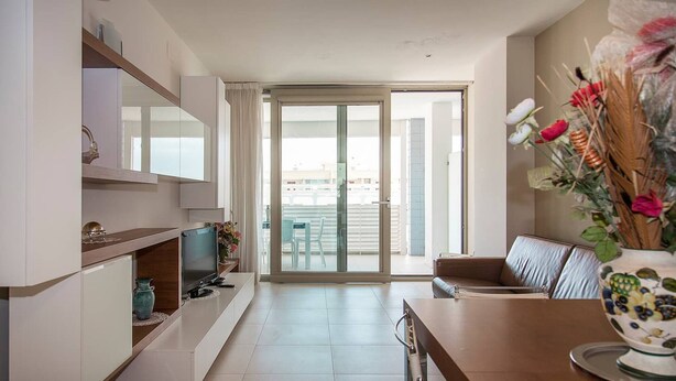 Uma sala de estar moderna com televisão de tela plana, sofá e mesa de jantar. Há uma porta de vidro deslizante que dá para uma varanda.