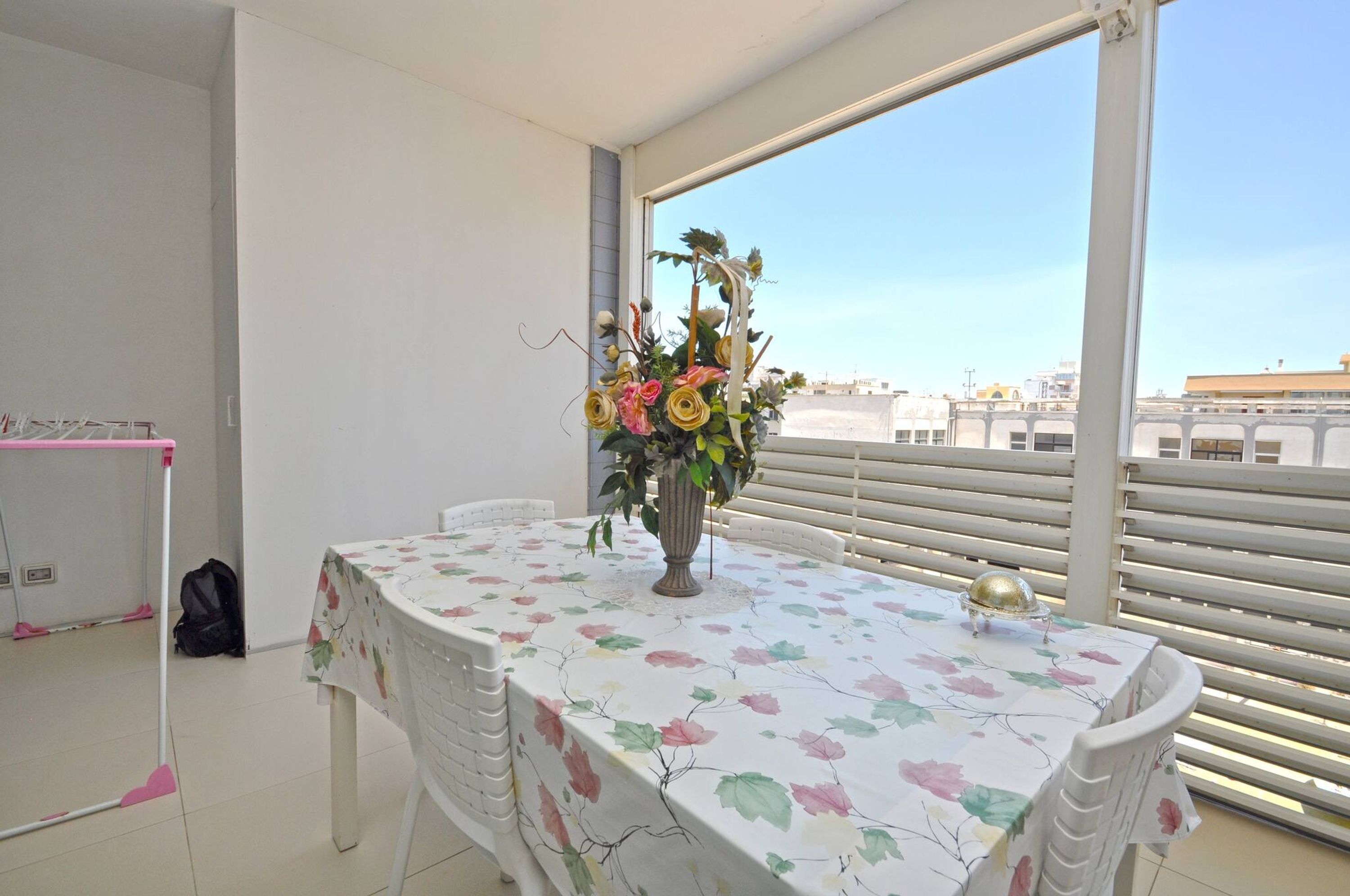 Een balkon met een tafel, afgedekt met een tafelkleed met bloemen, een vaas met bloemen en uitzicht op gebouwen.