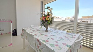 Uma varanda com uma mesa coberta por um tecido floral, um vaso com flores e vista para edifícios.
