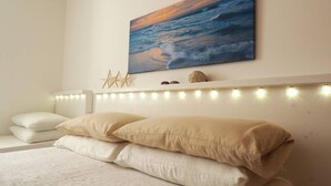 A bedroom with a bed, white pillows, a headboard with string lights, a painting of a sunset, and decorative shells. - Villa DEL Mare 2 in Torre San Giovanni (Ugento)