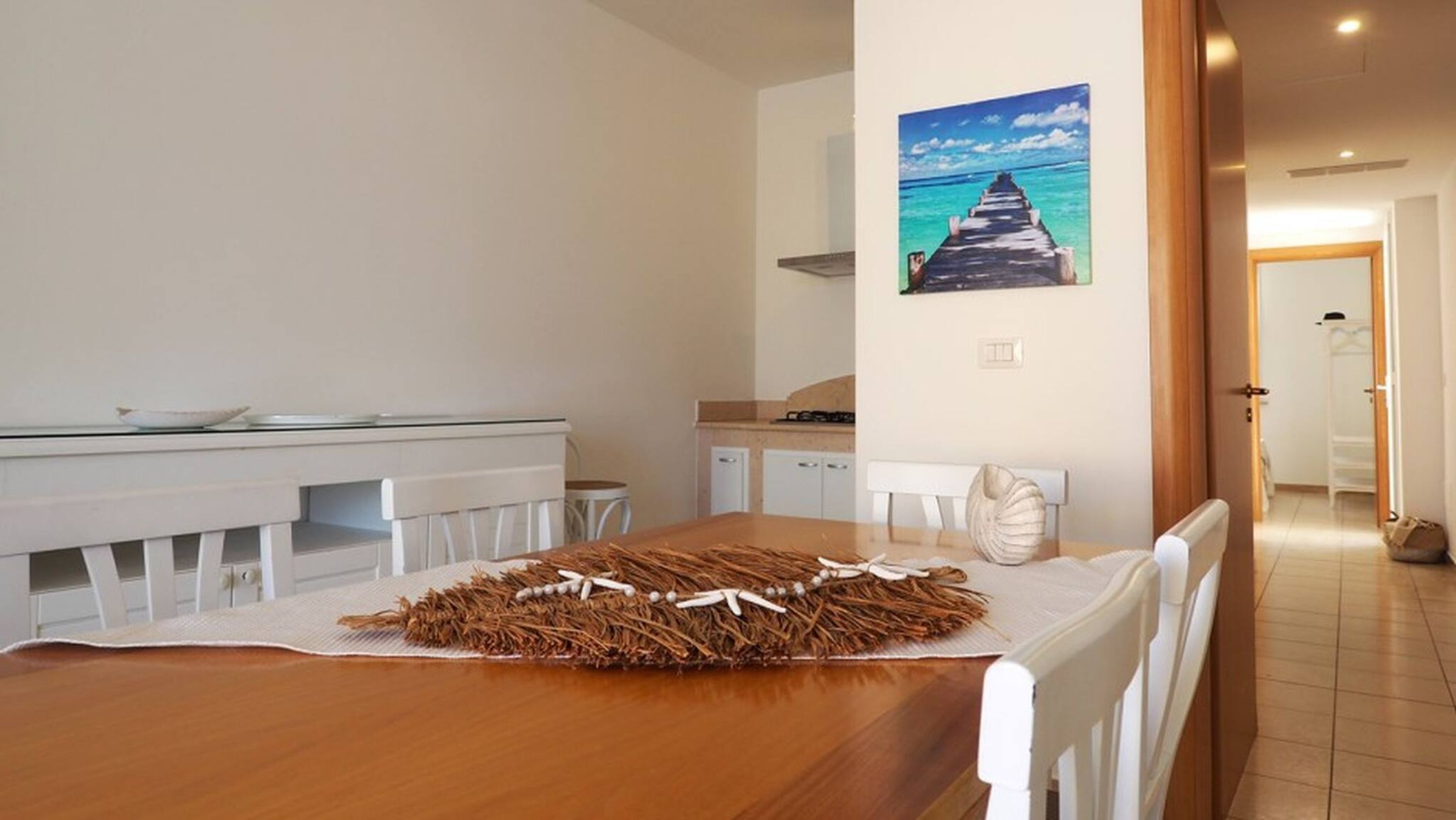 A dining area with a wooden table, white chairs, a white table runner, and a painting of a pier.