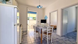 Une cuisine compacte comprenant un réfrigérateur, une table à manger avec des chaises, un téléviseur posé sur un meuble et un luminaire suspendu bleu.