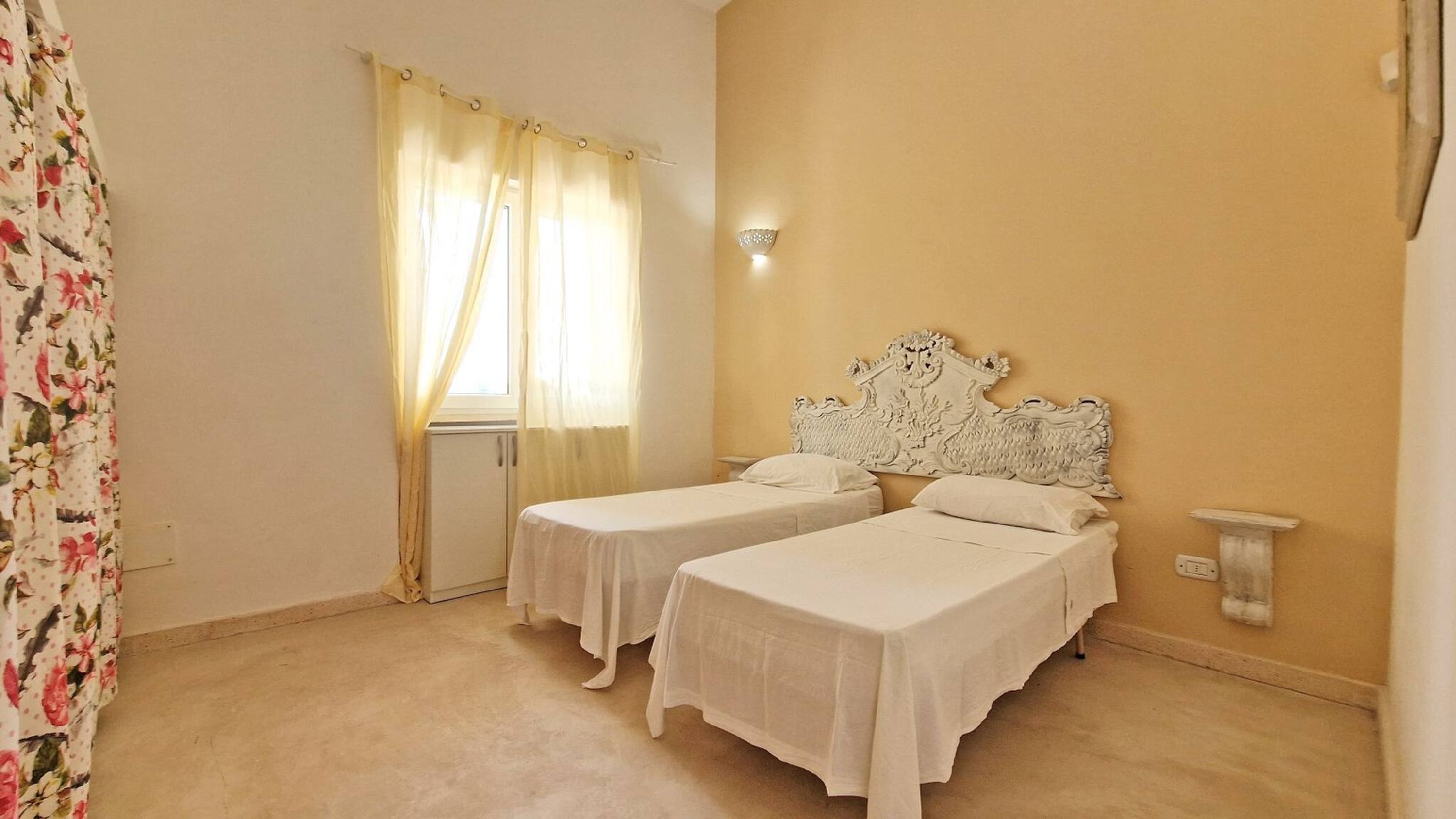 A room with two single beds, a window with sheer curtains, a wall-mounted light fixture, and a floral-patterned curtain on the left.