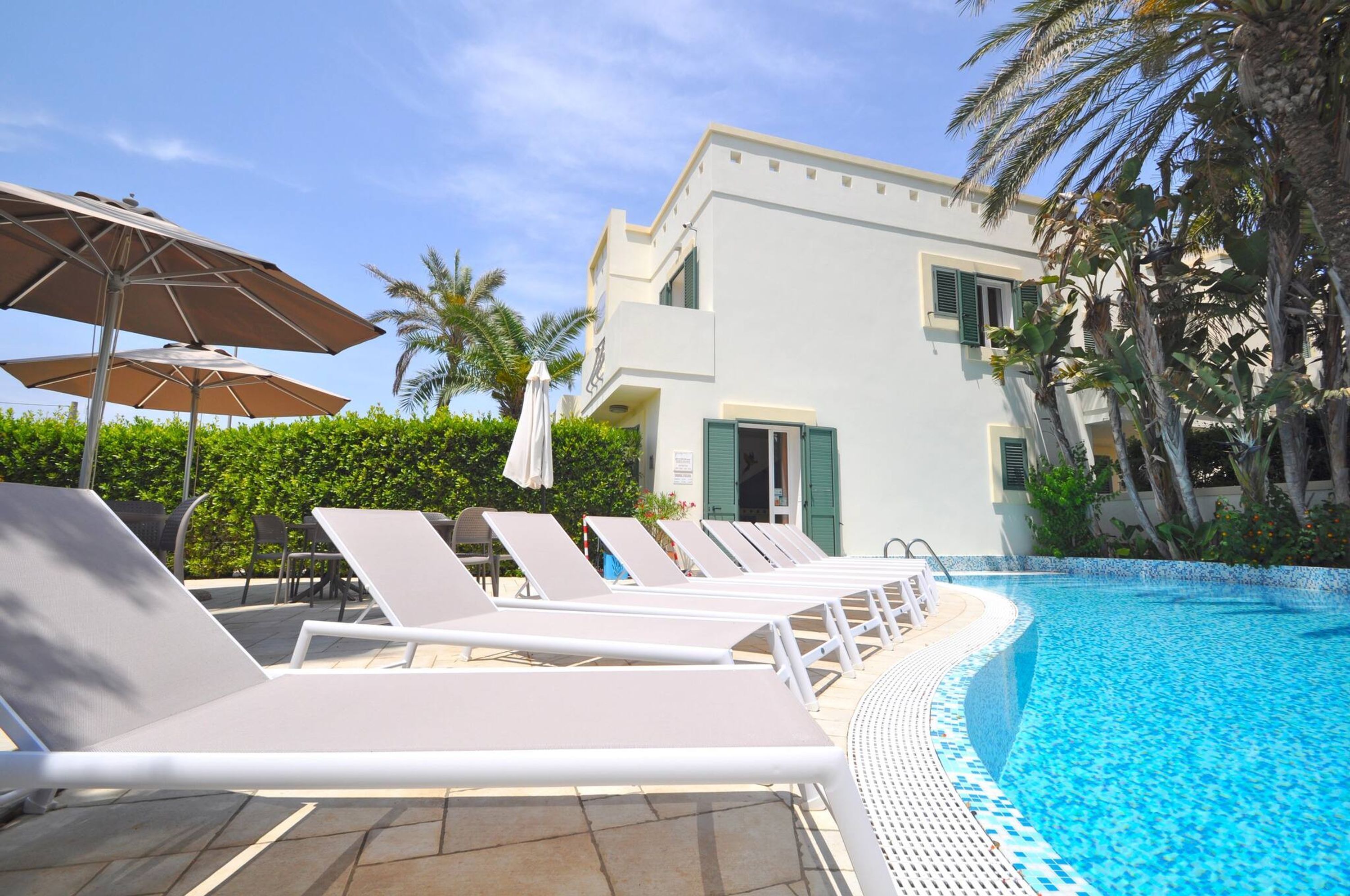 A poolside area with lounge chairs and umbrellas, a clear blue pool, and a white building with green shutters.