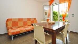 A dining area with a wooden table and four chairs, a patterned orange sofa, and a window with orange curtains. - Residence Arainn Kalepolis - Pool Sleeps 5 Wifi (Ugento)