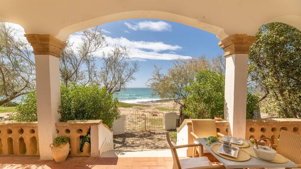 Une terrasse extérieure meublée de mobilier en osier, une table dressée avec une théière, des tasses et une assiette de nourriture, donnant sur une plage bordée par l’océan et des arbres.
