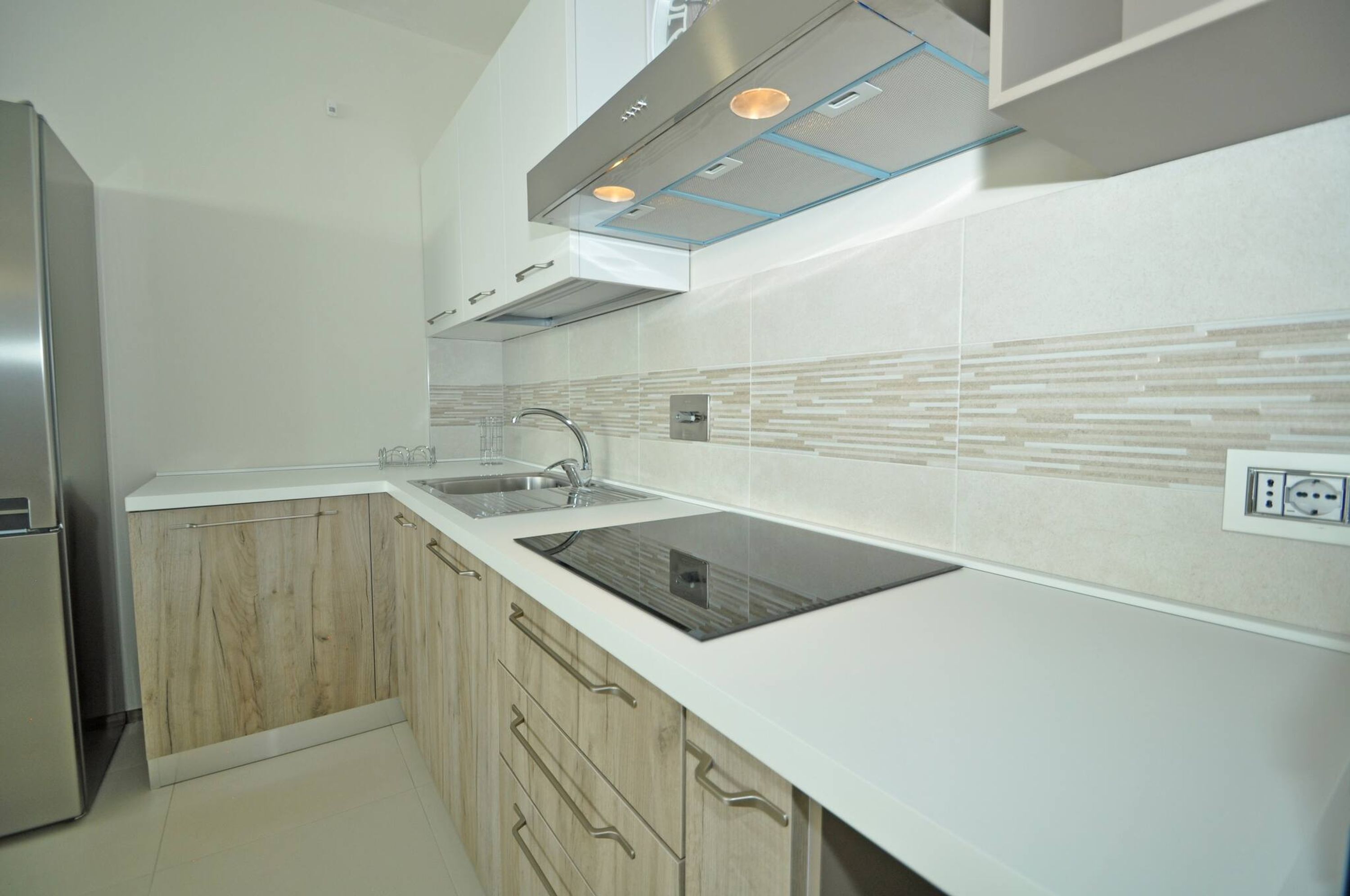 Una cocina moderna con un refrigerador de acero inoxidable, una placa de inducción negra, armarios de madera y una encimera blanca.